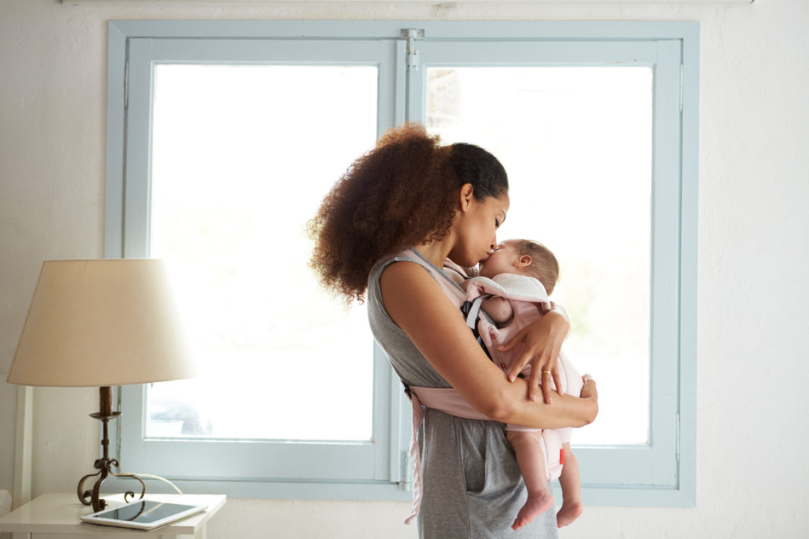 A woman cuddling a swaddled baby in front of a window.